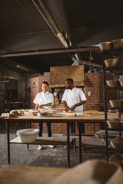 Multiracial male and female mid adult bakers talking while kneading dough at table in bakery
