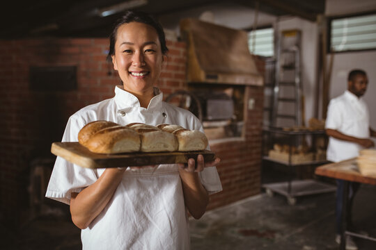 Portrait of smiling asian mid adult female baker holding serving board with fresh breads