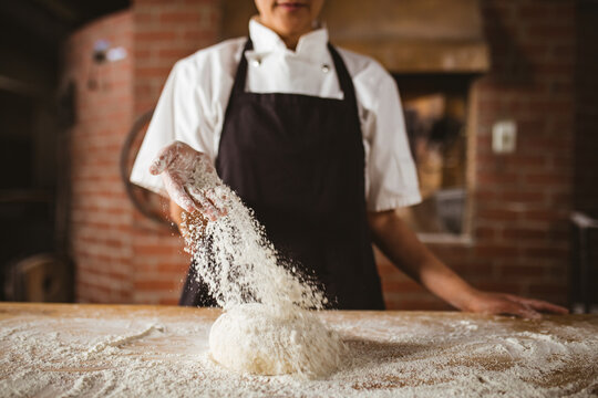 Midsection of asian mid adult female baker sprinkling flour on dough at table in bakery