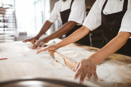 Midsection of multiracial mid adult female and male bakers rolling dough at table in bakery