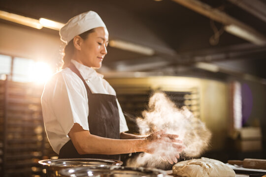 Asian mid adult female baker dusting flour on dough while working at table in bakery