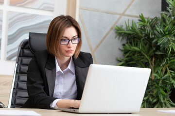 Portrait of successful and attractive business woman working on laptop in her office. Positive female employee in black suit smiling and sitting at the desk in her modern workplace