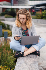 Woman student with curly hair and eyeglasses using laptop computer, mobile phone, studying, learning language. Communication, internet concept. Internet