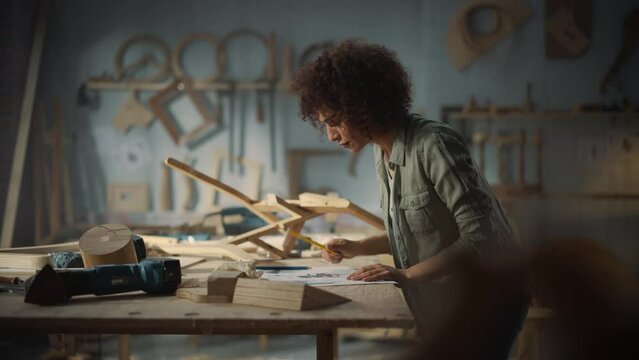 Portrait of a Young Beautiful Carpenter Checking a Blueprint and Starting to Assemble a Wooden Chair. Professional Furniture Designer Working in a Studio in Loft Space with Tools on the Walls.