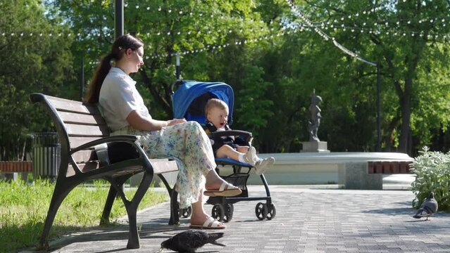 Mother with little cute child in baby stroller in the park, woman is resting on a bench on a sunny day.