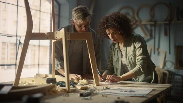 Portrait of Two Carpenters Working Together in a Woodwork Workshop. Multicultural Man and Female Colleagues Looking at a Blueprint on Paper and Discussing a New Chair Design.