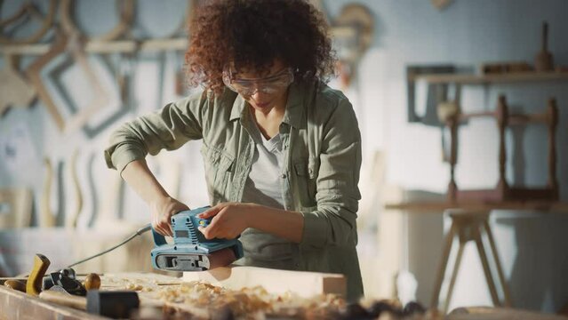 Multiethnic Woman Carpenter Wearing Protective Safety Glasses and Using Electric Belt Sander to Grind a Wood Block. Artist or Furniture Designer Working on a Product Idea in a Workshop.