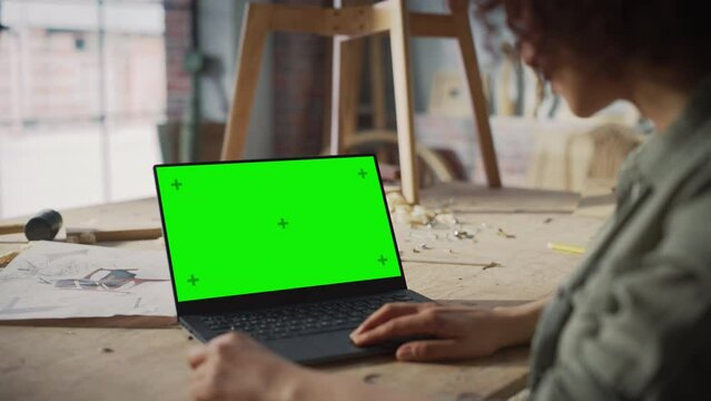 Over the Shoulder Shot of a Furniture Designer Working on Laptop Computer with Green Screen Mock Up Display. Creative Female Preparing a Design for Carpentry Project.