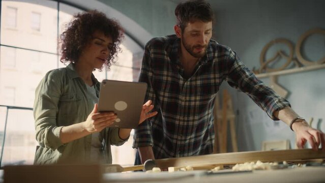 Small Business Owners of a Furniture Workshop Using Tablet Computer and Discussing the Design of a New Wooden Chair. Handsome Carpenter and Young Female Apprentice Working in Loft Studio.
