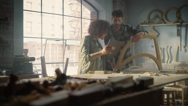 Portrait of Two Small Business Owners Using Tablet Computer and Discussing the Design of a New Wooden Chair in a Furniture Workshop. Carpenter and a Young Female Apprentice Working in Loft Studio.
