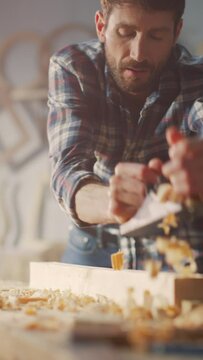 Vertical Screen: Handsome Young Artisan Craftsman in Checkered Shirt Using Hand Plane to Shape a Wood Bar. Carpenter Working on a Project in a Loft Studio with Tools on Walls.