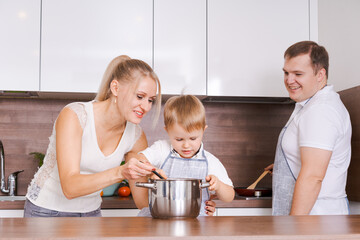 Obraz na płótnie Canvas Dad, mom and their little son are preparing dinner. Happy family preparing proper food in kitchen. Everyone smiles cheerfully together. Mom son stir soup in pan, dad fries vegetables in pan on stove