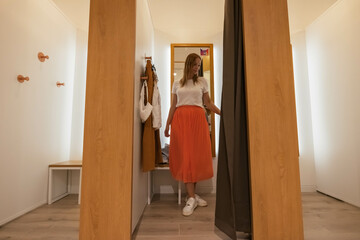 Fototapeta na wymiar a girl trying on a white t-shirt and an orange skirt in a booth looks in the mirror. brown and white fitting booths in a things store. there is a place for an inscription