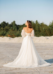 A pretty brunette girl in a white dress stands casually with a bouquet of dry flowers on the background of a white sand quarry, wedding concept