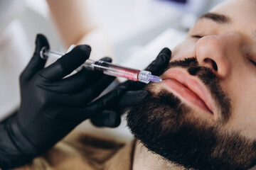 Stylish bearded man visiting aesthetic clinic, getting lips filler, closeup. Attractive Man having beauty injection at male spa salon. Anti-aging treatment for men concept.