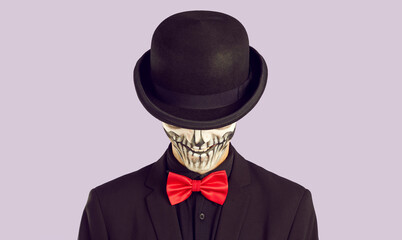 Studio shot of skeleton man in stylish Halloween outfit. Portrait of man with skull makeup dressed in black suit and red bow tie hiding his face under black hat isolated on light purple background