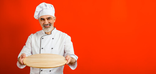 Chef-cooker in a chef's hat and jacket holding an empty wooden tray for mock up. Senior professional baker man wearing a chef's outfit. Character kitchener, pastry chef for advertising