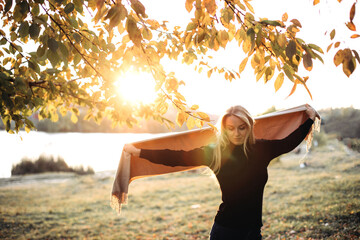 Beautiful girl with long blond hair in autumn landscape at sunset. Autumn portrait of a girl holding a scarf, front view, selective focus