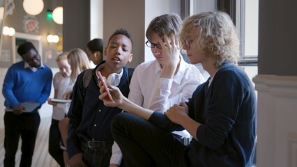 Multiracial young friends using mobile phone while sitting on windowsill in school corridor