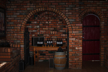 Vaulted red brick wine cellar at the winery. Old oak barrel and a stack of bottles against dark...