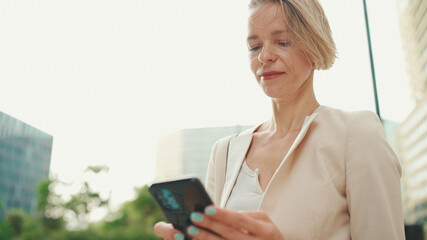 Smiling woman with blond hair wearing beige suit using cellphone while sitting outside on the street