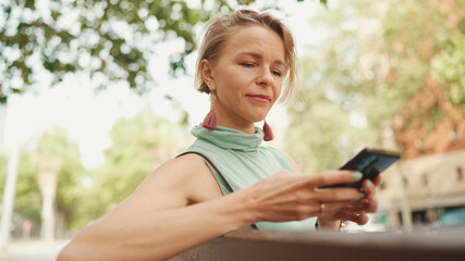 Beautiful happy smiling woman with short blond hair in casual clothes sits on bench, sends voice message on cellphone