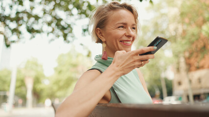 Beautiful happy smiling woman with short blond hair in casual clothes sits on bench, sends voice message on cellphone