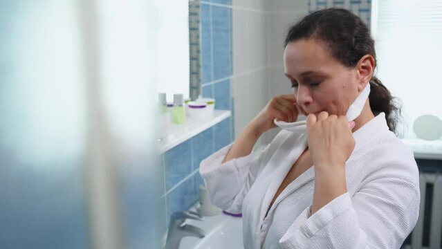 Beautiful middle-aged multi-ethnic woman wearing a white bathrobe, stands in the bathroom and puts on a terry headband before morning beauty procedures. Hygiene. Skin and body care. Daily routine
