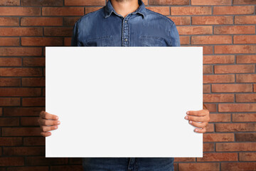 Man holding white blank poster near red brick wall, closeup. Mockup for design