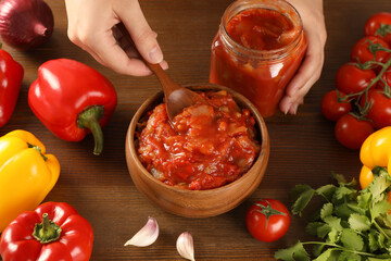 Woman putting tasty lecho from jar into bowl at wooden table, above view