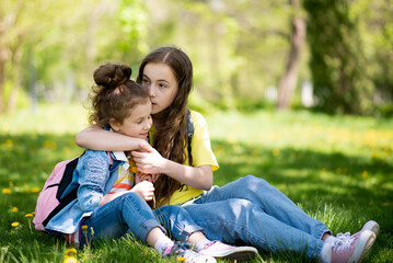 Naklejka na ściany i meble Two cute sisters' girls on a walk. It's spring outside and the sun is bright. A girl holds a dandelion in her hands. She is wearing a colored T-shirt and a denim shirt. Childhood. Family.