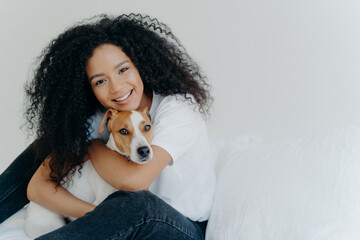Photo of attractive young woman with Afro hircut, embraces with love dog, takes care of pet, smiles gently, wears casual clothing, isolated over white background, sit on bed, copy space for your promo