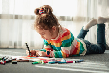 A cute little girl sits on the floor in the rays of the sun and draws a rainbow with colorful markers. She is dressed in a colored jumper. Creation. Childhood.