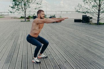 Full length shot of active man does squatting exercise poses with naked torso has muscular body...