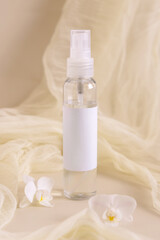 Fototapeta na wymiar Spray refillable bottle near orchid flowers and tulle on light beige. Cosmetic packaging Mockup