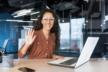 Fototapeta na wymiar Portrait of young successful business woman in modern office using laptop, Hispanic woman with curly hair smiling and looking at camera, holding hand up in greeting gesture.