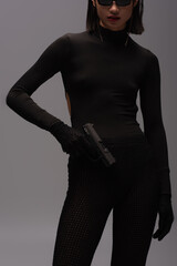 cropped view of dangerous asian woman in total black outfit and stylish sunglasses holding gun isolated on grey.