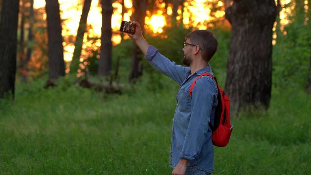 Male tourist with backpack make photos with mobile phone in summer landscape in forest at sunset. Travel blogger shoot video