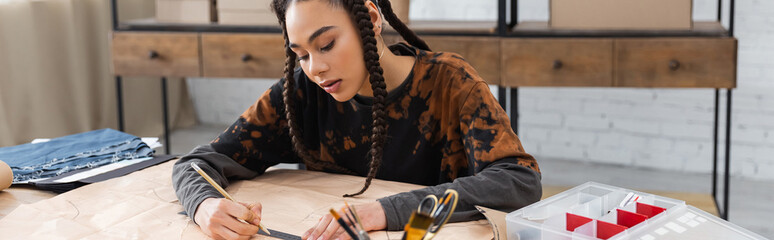 African american designer drawing on sewing pattern near cloth and decor in workshop, banner.