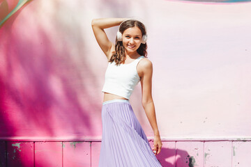 a young excited woman in a top and skirt, wearing headphones, listening to music, walking, dancing with her hands up in the open air against a pink wall.