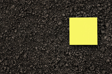 Yellow note paper square on black soil texture ground close up. Yellow sticky note blank paper flat...