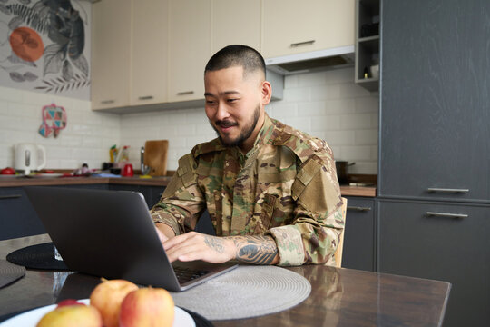 Satisfied military asian sits at table and works on laptop