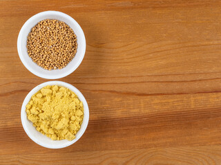 brown Mustard seeds and yellow mustard powder in white bowls on a wooden table
