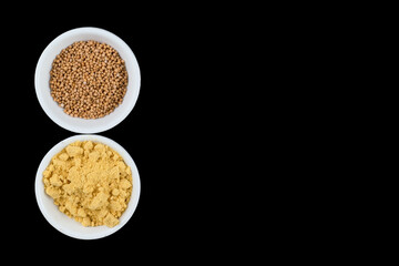 brown Mustard seeds and yellow mustard powder in white bowls on a black background