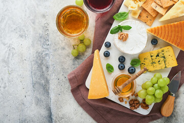 Assortment of cheese, honey, cracker, blueberries, grapes with red and white wine in glasses antipasto server on white marble board on grey background. Flat lay, copy space.