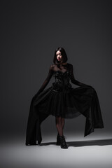 full length of asian woman in black gothic dress and stylish boots posing on dark grey.