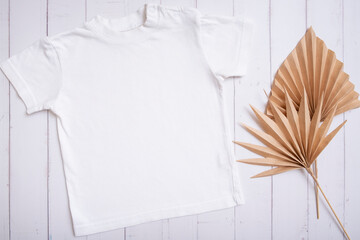 White children's t-shirt mockup for logo, text or design on wooden background with palm leaves top view