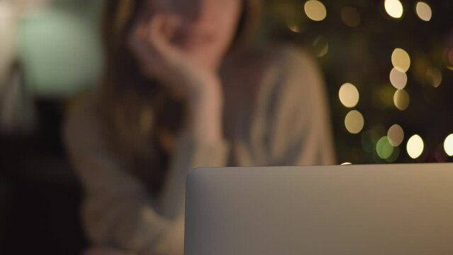 4K video. Girl freelancer in knitted sweater working at home with laptop notebook. Bokeh of festive lights of Christmas tree. Chatting on computer, surfing internet, shopping online selective focus