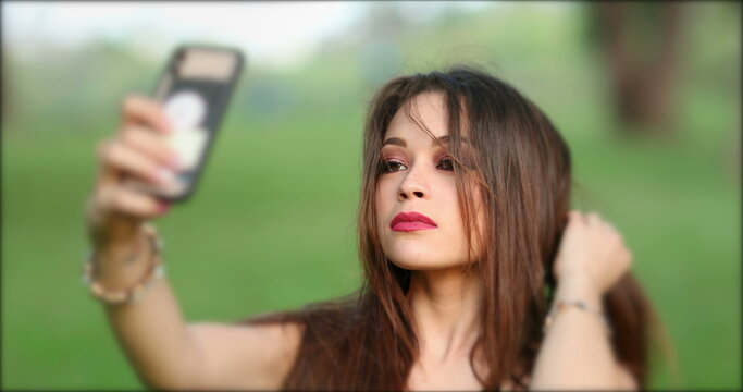 Pretty young woman taking selfie with smartphone device. Girl adjusting hair preparing to take photo of herself with phone
