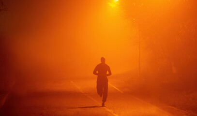 Silhouette of a runner in the fog. He runs in the evening urban environment in the fog, which is highlighted by the warm light of lanterns.